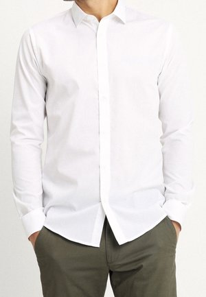 Homme portant une chemise blanche ajustée à manches longues avec col et boutons, et un pantalon vert olive, mains dans les poches, visage non visible.