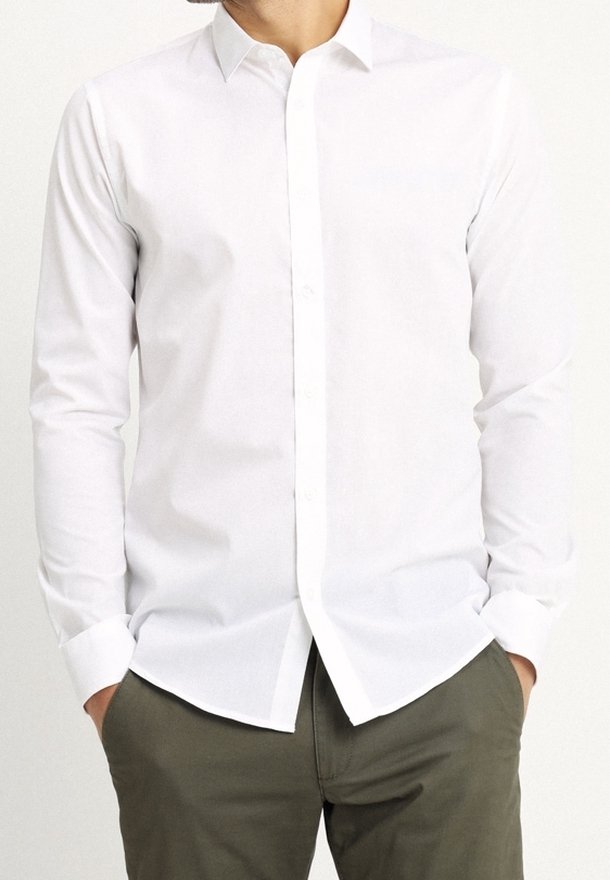 Homme portant une chemise blanche ajustée à manches longues avec col et boutons, et un pantalon vert olive, mains dans les poches, visage non visible.
