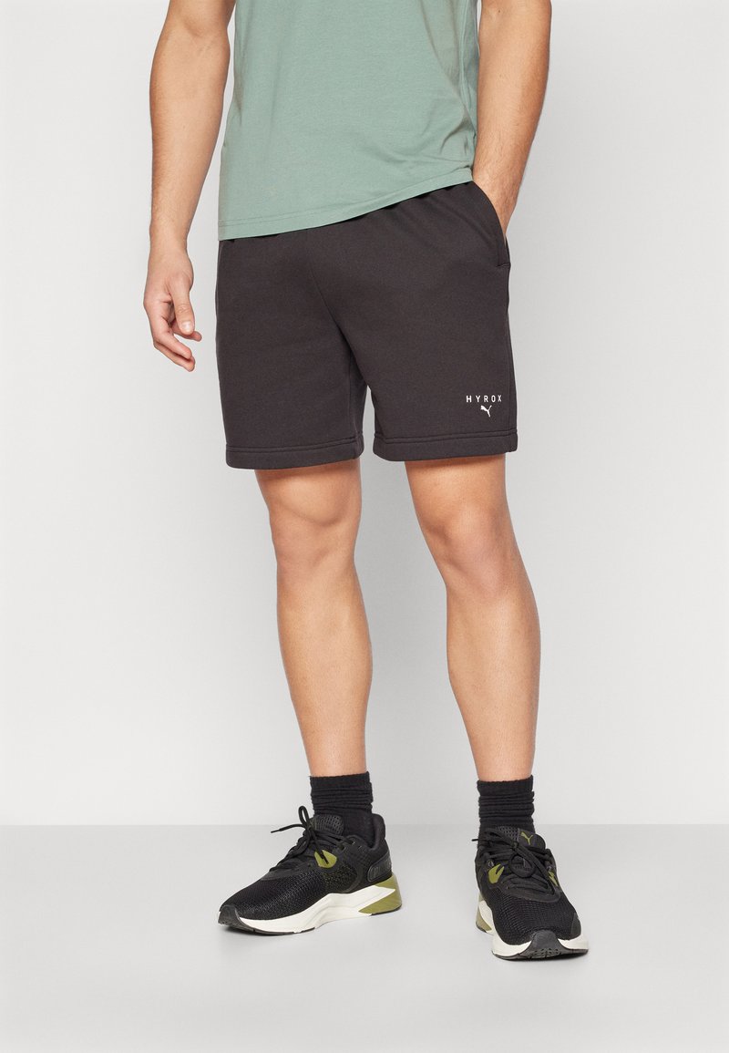Shorts de sport gris foncé avec une taille élastique, des poches latérales et un logo blanc au bas. Associés à des baskets noires et des chaussettes.