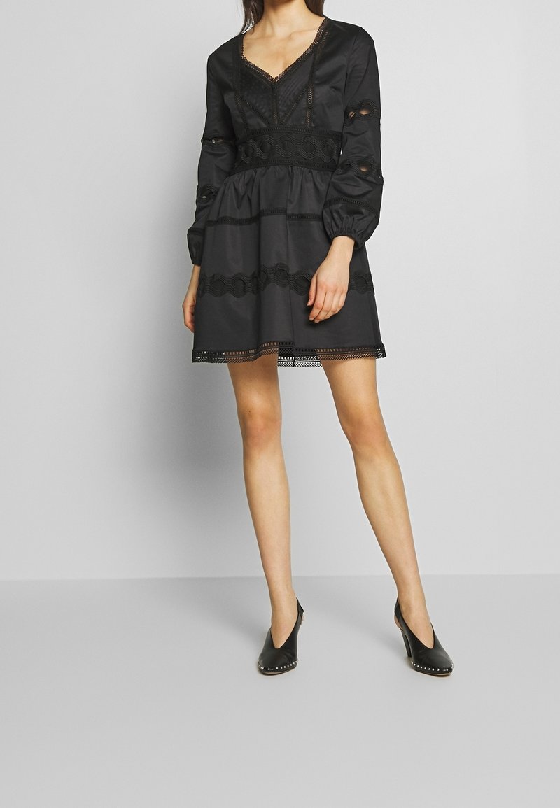 Robe noire à manches longues, col en V, avec des détails en dentelle à la taille et à l'ourlet. Elle présente une jupe évasée et des découpes sur les manches.
