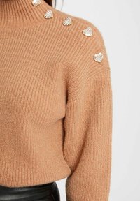 Pull en tricot épais beige avec une texture côtelée. Présente des ornements en forme de cœur argenté sur l'épaule.
