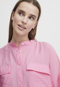 Camicia button-up a maniche lunghe di colore rosa brillante. Presenta un colletto alla cinese, due tasche frontali e una texture stropicciata con una vestibilità comoda.