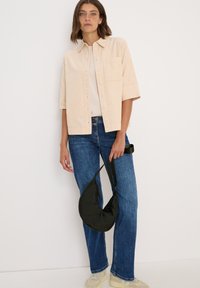 Ljusbeige skjorta med korta ärmar och bröstficka, i kombination med blå jeans och en mörkgrön väska. Sneakers fulländar outfiten.