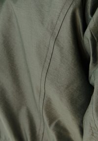 Tkanina oliwkowo-zielona o gładkiej fakturze, z delikatnym połyskiem i drobnym przeszyciem wzdłuż krawędzi. Brak widocznych wzorów lub elementów metalowych.