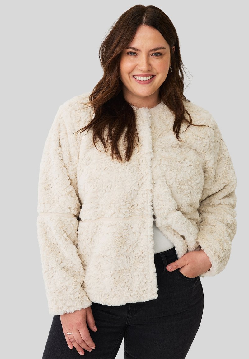 Lachende vrouw met lang bruin haar, gekleed in een crème-kleurige fluffy jas, witte top en zwarte broek, met een hand in haar zak op een grijze achtergrond.