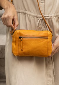 Person som bär en beige klänning och håller en liten gul läder crossover-väska med ficka med dragkedja fram och justerbar rem.