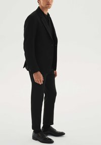 Fekete, testhez álló szabású öltönykabát és nadrág, fekete bőrcipővel párosítva. Simak, vékony szabású, minimalista stílus, minták nélkül.