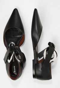 Zapatos de cuero negro de punta afilada con tacón plano, que presentan un diseño recortado y un acento circular plateado en la correa del tobillo.