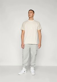 Muž stojící v béžové košili s krátkým rukávem, šedých cargo teplákách a bílých vysokých teniskách na jednobarevném pozadí.