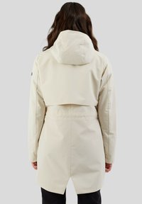 Veste imperméable beige avec une capuche ajustée, une taille cintrée et un ourlet arrière plus long. Texture lisse avec un minimum de quincaillerie visible.