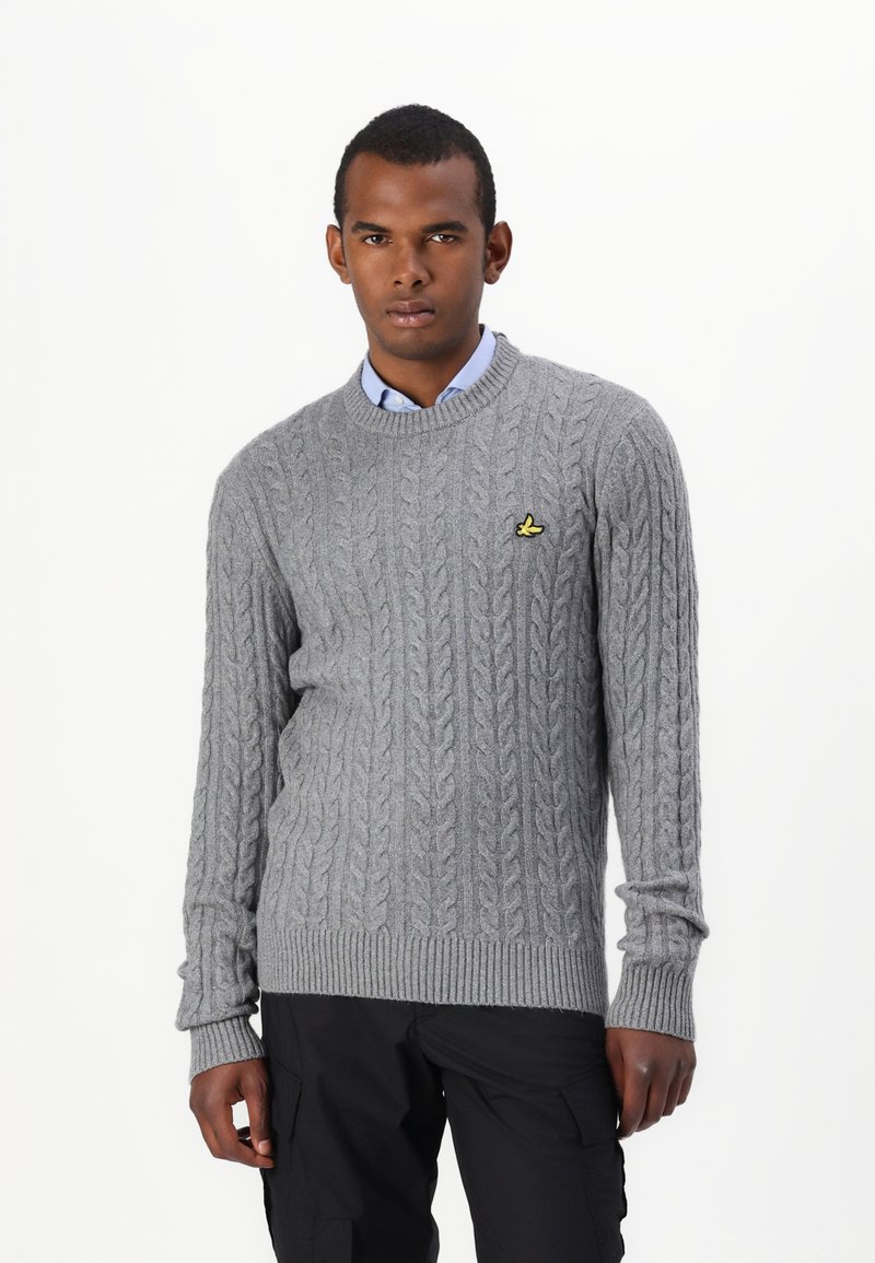 Suéter de punto grueso gris claro con cuello redondo y puños acanalados, que incluye un pequeño logo amarillo en el lado izquierdo del pecho.