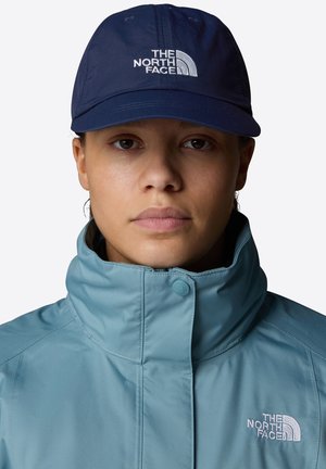 Light blue waterproof jacket with high collar and embroidered logo, paired with a dark navy cap featuring a white logo.