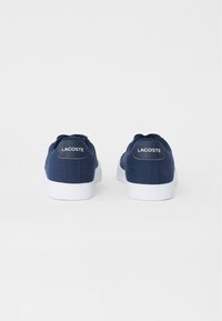 Zapatillas de lona azul marino con suelas de goma blanca. Presentan una puntera redondeada y un logo de Lacoste en el talón trasero. Diseño casual, perfil bajo.