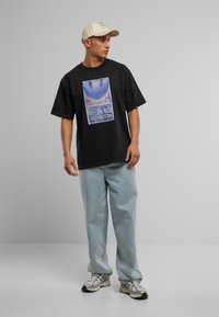 Svart oversized t-skjorte med et fargerikt grafisk trykk, sammen med lyseblå baggy jeans og grå joggesko. Lysbeige caps brukt.