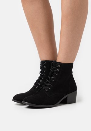 Botas de tornozelo em camurça preta com bico arredondado, atacadores à frente e um salto em bloco empilhado. O design apresenta ilhós metálicos para os atacadores.
