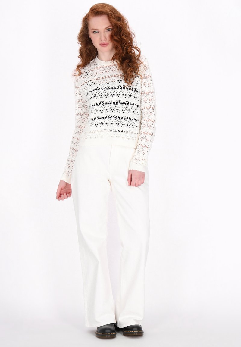 Cropped sweter z koronkowym splotem w kolorze kremowym z czarnymi poziomymi paskami. Połączony z białymi spodniami o wysokim stanie i szerokimi nogawkami oraz czarnym obuwiem.