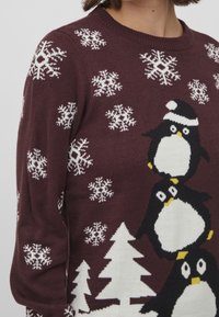 Vinröd tröja med tre pingviner staplade, med vita snöflingor och träd i ett stickat mönster. Mjuk textur, rund halsringning.