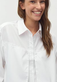 Camicia bianca con colletto classico, caratterizzata da una texture liscia e due tasche frontali con chiusura a bottone.