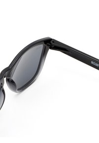 Mustat aurinkolasit, joissa on paksu, kiiltävä muovirunko, tummennetut linssit ja ohuet metalliset sangoissa logo-yksityiskohta.