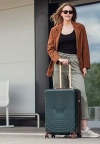 Zelený tvrzený kufr s vlnkovým vzorem, hnědými akcenty, výsuvnou rukojetí a čtyřmi kolečky. V doprovodu osoby v hnědém blejzru.