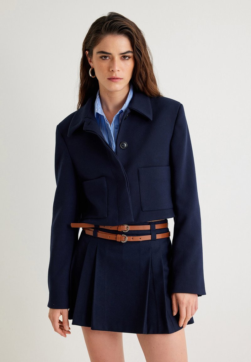 Granatowa krótka kurtka z dużymi klapami, zapięciem na guziki i kołnierzem, noszona na niebieskiej koszuli w paski oraz plisowanej spódnicy z brązowym paskiem.