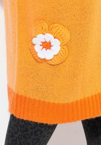 Pletený oranžový svetr s oranžovým žebrovaným lemem, se sladěnou háčkovanou květinou v oranžové a bílé na přední straně. Viditelný detail texturovaného materiálu.
