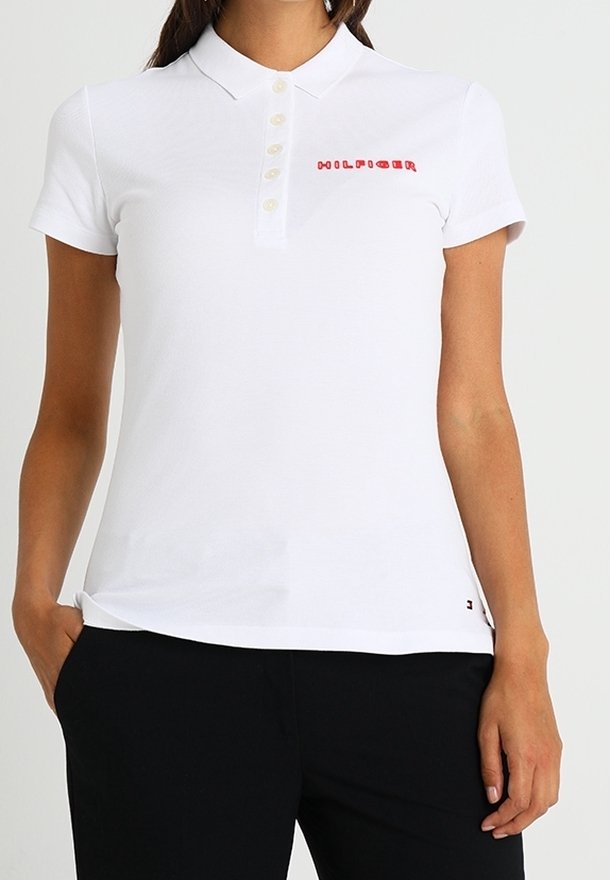 Femme portant un polo blanc à manches courtes avec cinq boutons et un texte rouge sur la poitrine, associé à un pantalon noir.