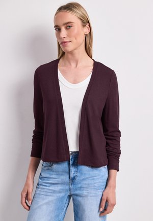 Lyshåret kvinde iført en mørke lilla åben front cardigan over en hvid top og lyseblå jeans mod en ensartet lys baggrund.