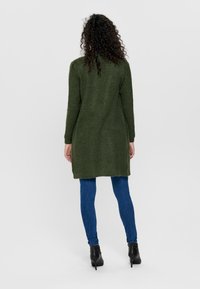 Donkergroene gebreide lange cardigan met geribbelde textuur, gecombineerd met blauwe skinny jeans en zwarte enkellaarzen, gezien vanaf de achterkant.