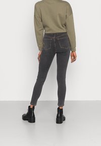 Jeans ajustados de mezclilla gris oscuro con cintura alta, diseño de cinco bolsillos y costuras visibles. Combinados con un sudadera verde holgada y botas negras.