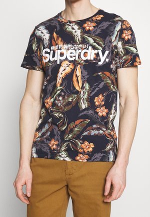 Mann trägt ein dunkles T-Shirt mit Blumenmuster, „Superdry“-Logo und japanischem Text, kombiniert mit senfbraunen Hosen, steht vor einfachem Hintergrund.