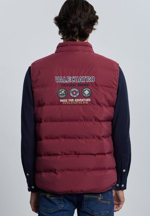 Persona que lleva un chaleco acolchado granate con "VALECUATRO Made for Adventure" y logotipos en la espalda sobre una camisa de manga larga oscura.