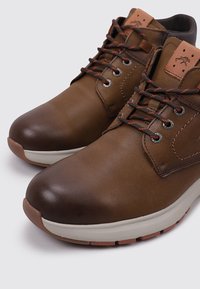Bottines en cuir marron avec finition texturée, bout arrondi, œillets en métal et lacets orange. Semelle extérieure beige avec motifs de grip.