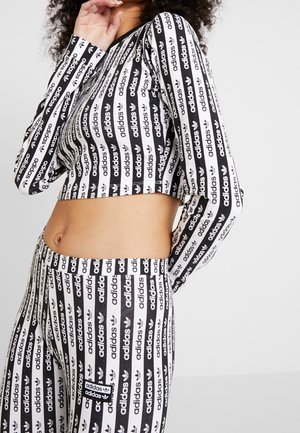 Kvinde iført en sort-hvid Adidas crop top og matchende bukser med gentagne Adidas-logoer i lodrette striber, der viser hendes mave.