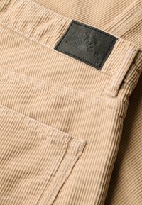 Pantalon en velours côtelé beige avec une poche arrière et un patch rectangulaire noir affichant "Tokyo 1967" et un logo du soleil levant.