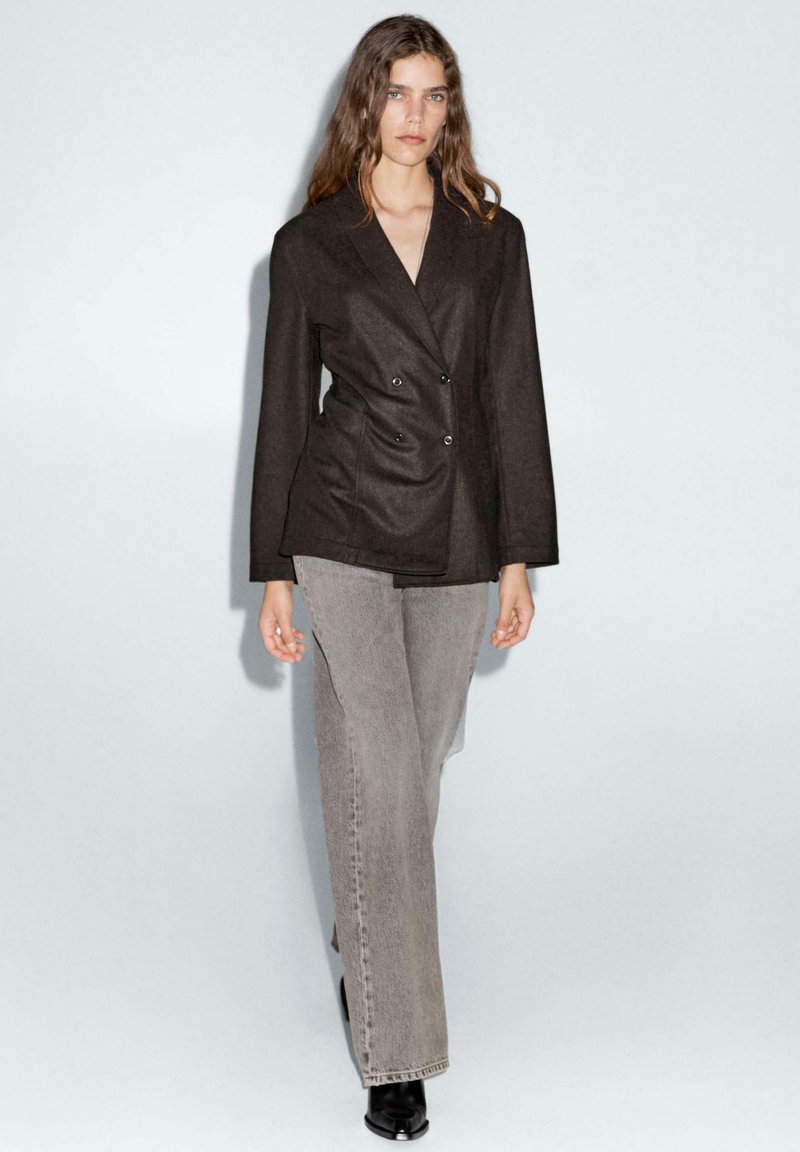 Blazer noir à double boutonnage avec des revers structurés, associé à un jean large gris clair et des bottines noires. Texture de tissu lisse.