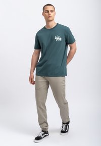 Turquoise katoenen t-shirt met korte mouwen en een bedrukte afbeelding op de linkerkant van de borst, gecombineerd met lichtbeige tapered broeken en zwarte sneakers.