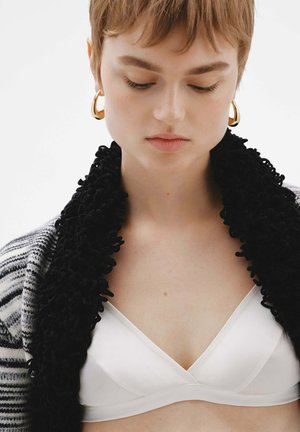 Joven con el cabello corto que lleva pendientes de aro dorados, una bufanda negra texturizada, un suéter de rayas blanco y negro y una blusa blanca, mirando hacia abajo.
