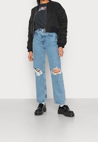 Svart bomberjacka, marinblå grafisk t-shirt, ljusblå slitna vida jeans och svarta plattformsskor med strukturerad sula.