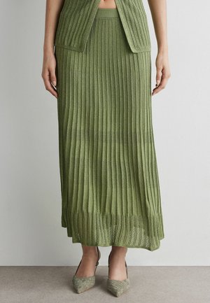 Kobieta nosząca midi spódnicę z oliwkowozielonego prążkowanego dzianego materiału, dopasowany top oraz szpiczaste plecione buty typu slingback.