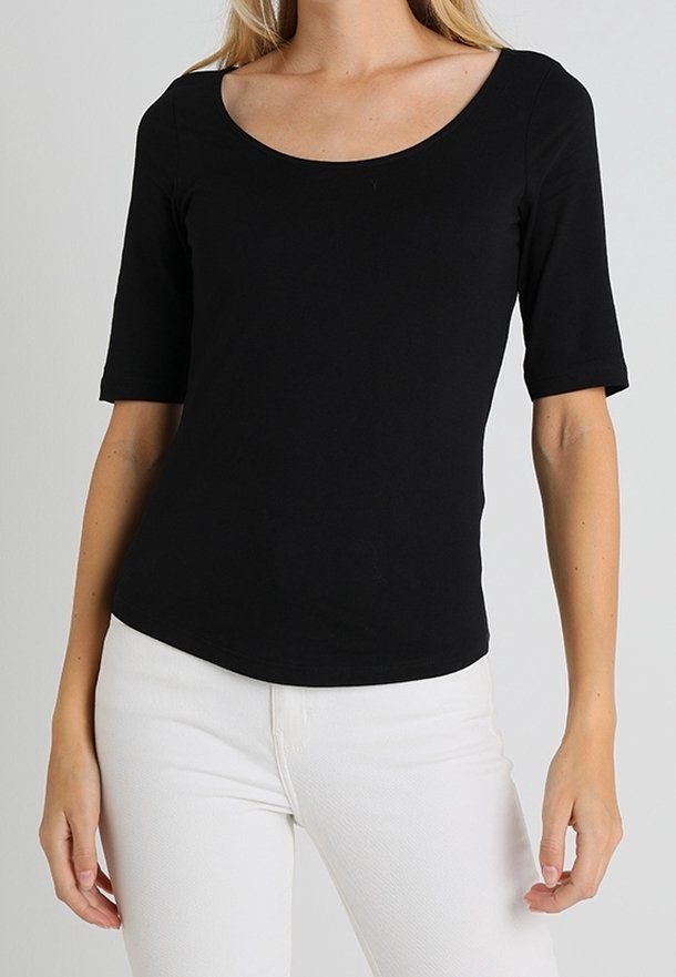 Femme portant un haut noir ajusté à manches mi-longues avec un col rond et un pantalon blanc sur un fond uni.