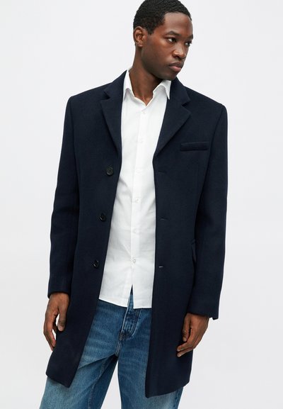 Navy wool overcoat featuring a notched collar, three front buttons, and a chest pocket; paired with a white shirt and blue jeans.