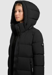 Doudoune noire avec une grande capuche, fermeture éclair à l'avant et poches latérales. Le tissu texturé présente des coutures horizontales et un patch logo.