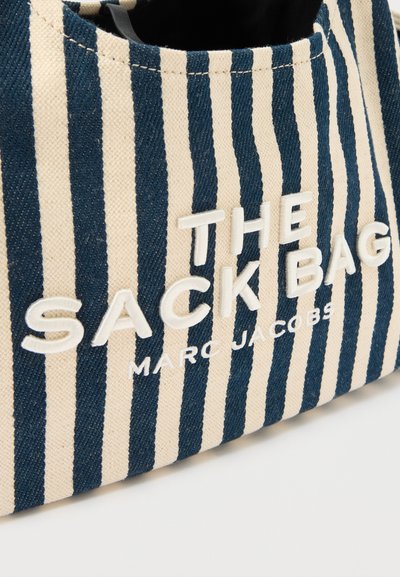 Τσάντα από ύφασμα με ναυτικό μπλε και εκρού ρίγες, με ανάγλυφα λευκά γράμματα που αναγράφουν «THE SACK BAG MARC JACOBS» σε απλή επιφάνεια.