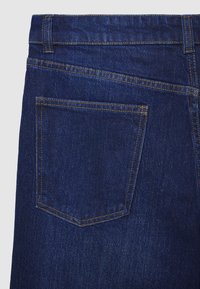 Retro di tasca posteriore di jeans in denim blu scuro con cuciture arancioni e passante della cintura su uno sfondo chiaro.