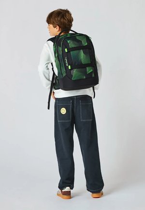 Child facing away, wearing black pants with smiley patch, white sweatshirt, brown Adidas shoes, and green-black geometric patterned backpack.