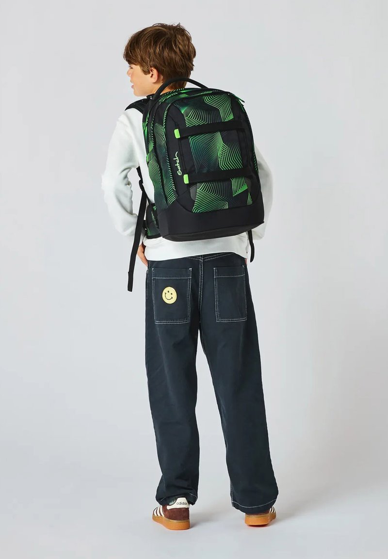 Kind mit dem Rücken zur Kamera, trägt schwarze Hose mit Smiley-Aufnäher, weißen Pullover, braune Adidas-Schuhe und einen Rucksack mit grün-schwarzem geometrischem Muster.