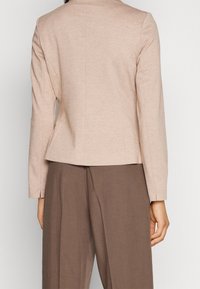 Blazer beige sur mesure à manches longues, design structuré, pinces au dos et texture subtile, associé à un pantalon marron.