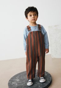 Brune og sorte stribede overalls lavet af denim, parret med en lys blå langærmet skjorte og sorte sneakers med hvide såler.