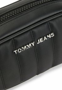 Black leather bag with a quilted design, silver "TOMMY JEANS" logo, and a top zipper. Features textured surface and detailed stitching.